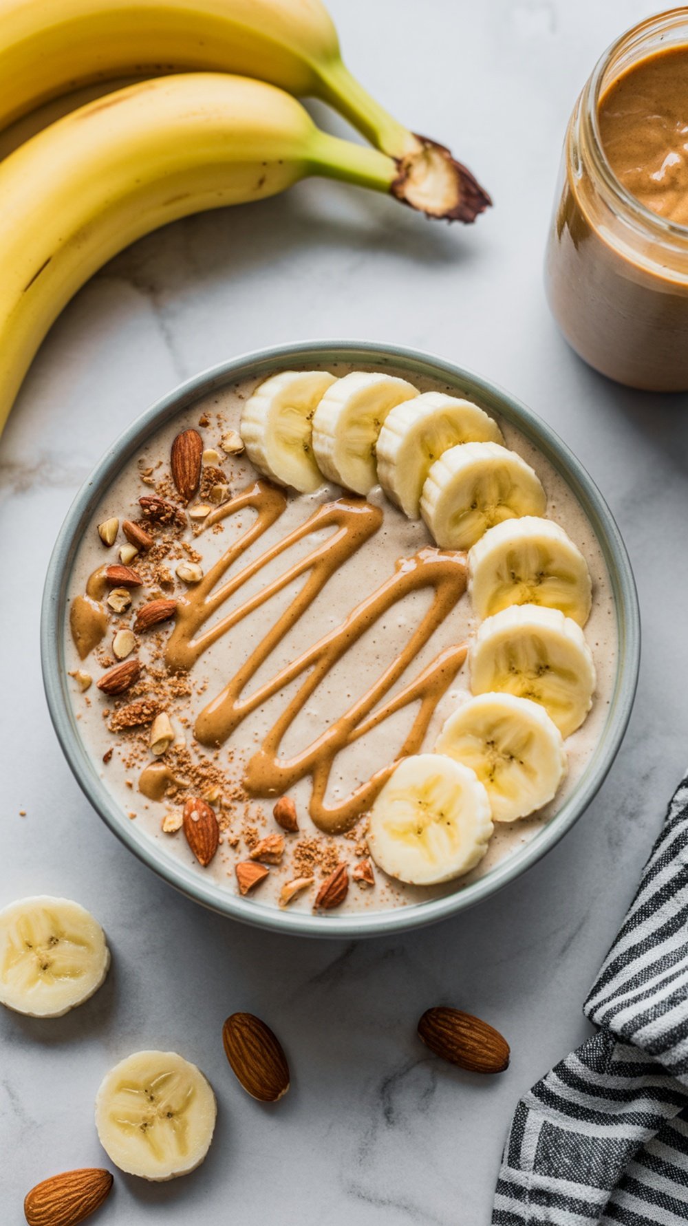 A bowl of banana and almond butter smoothie topped with banana slices, almonds, and almond butter, with bananas in the background.